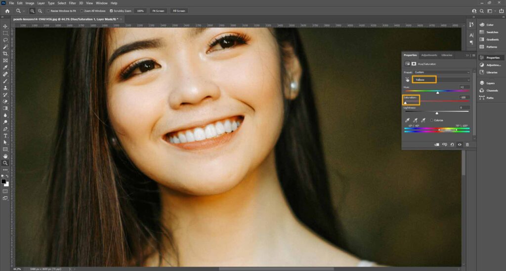 A woman smiling while using Adobe Photoshop, demonstrating a Hue/Saturation adjustment technique on teeth.
