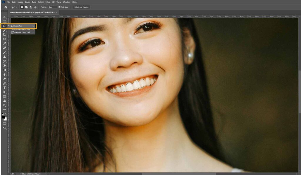A woman smiling while using Adobe Photoshop, demonstrating the Lasso tool to select the teeth.