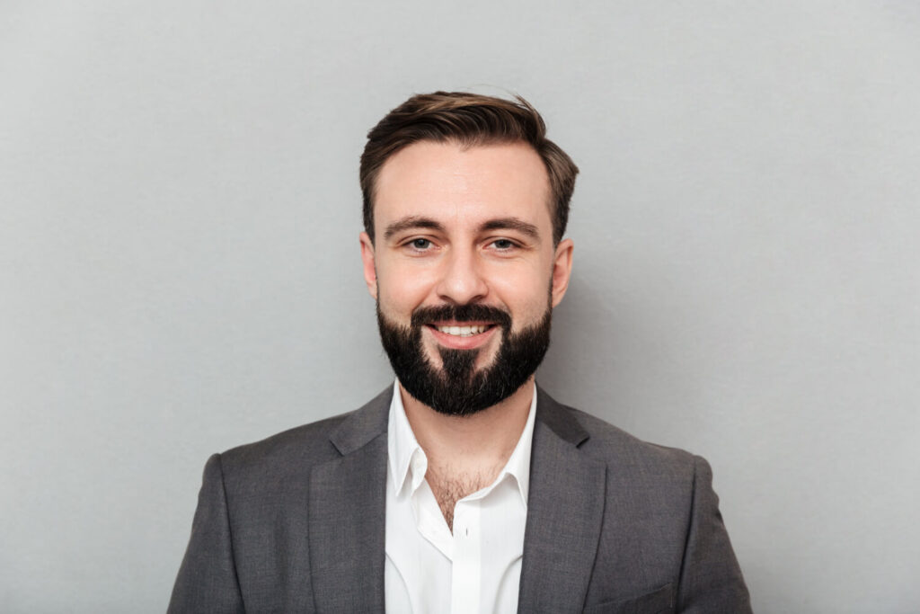 A handsome young man with a beard, wearing a white shirt against a grey background, suitable for corporate headshots.