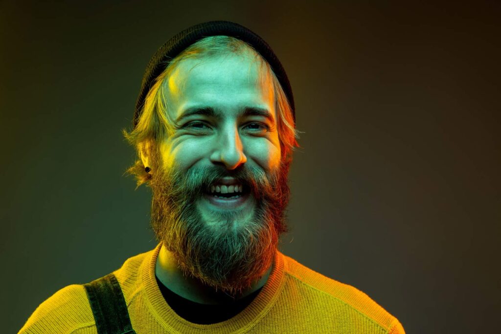 A smiling man with a beard and a hat poses for a creative headshot.
