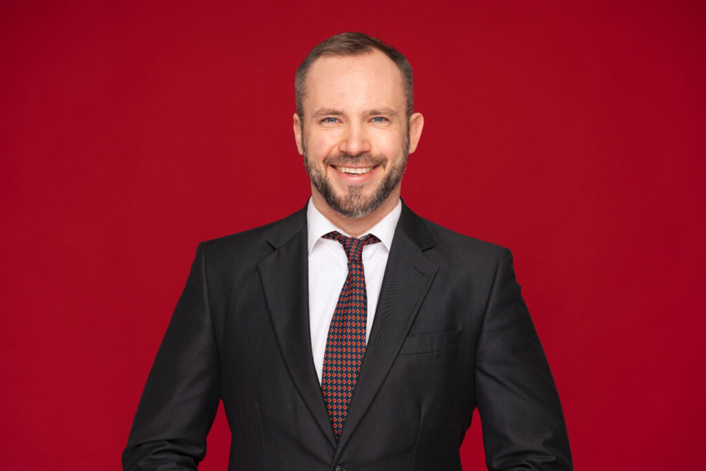 A man in a suit and tie poses against a simple red background, showcasing a simple headshot photography background.