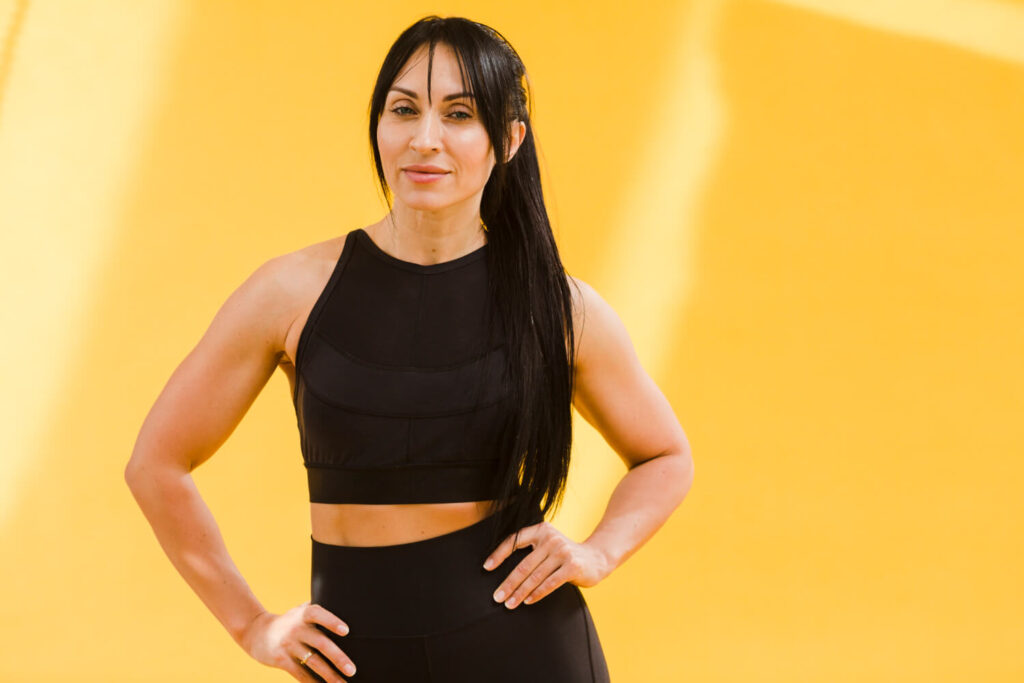 A woman in black sports gear poses confidently against a bright yellow background, showcasing a fitness headshot.