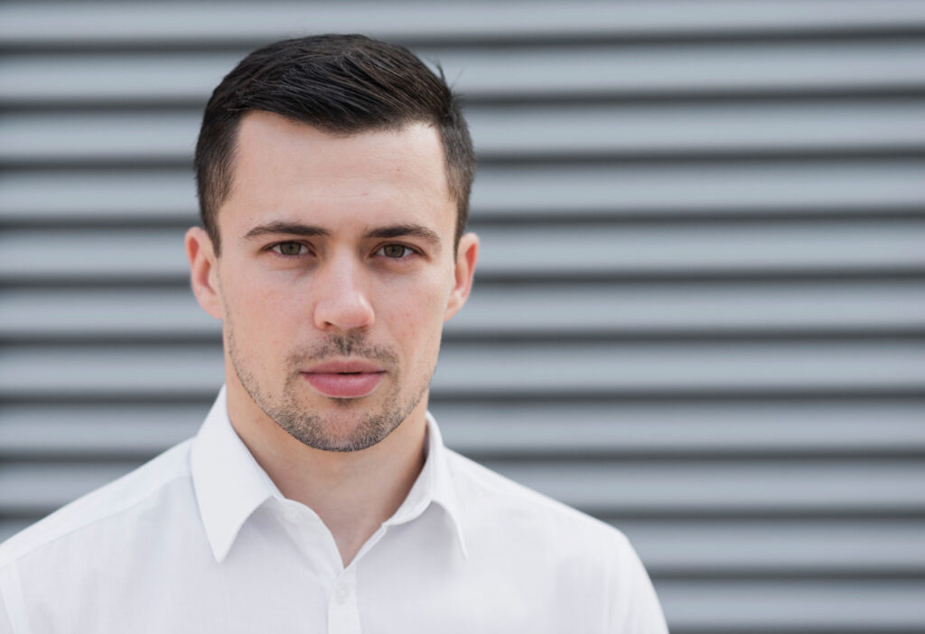 Headshot of a man wearing a white shirt, standing confidently in front of a neutral wall background.