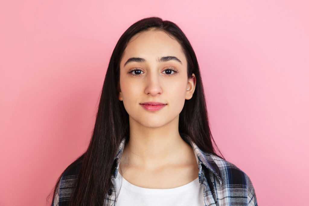 Headshot of a young woman with long hair, set against a soft pink background, emphasizing feminine beauty.