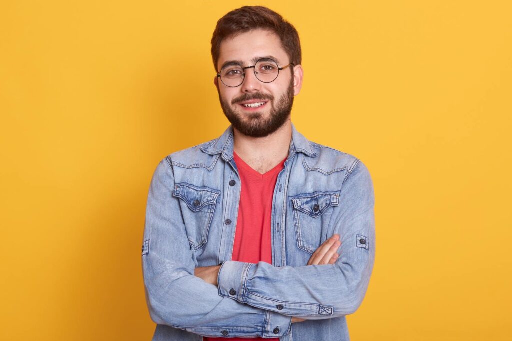 Young man in glasses and a denim jacket poses against a bright yellow background, showcasing a high-quality headshot.