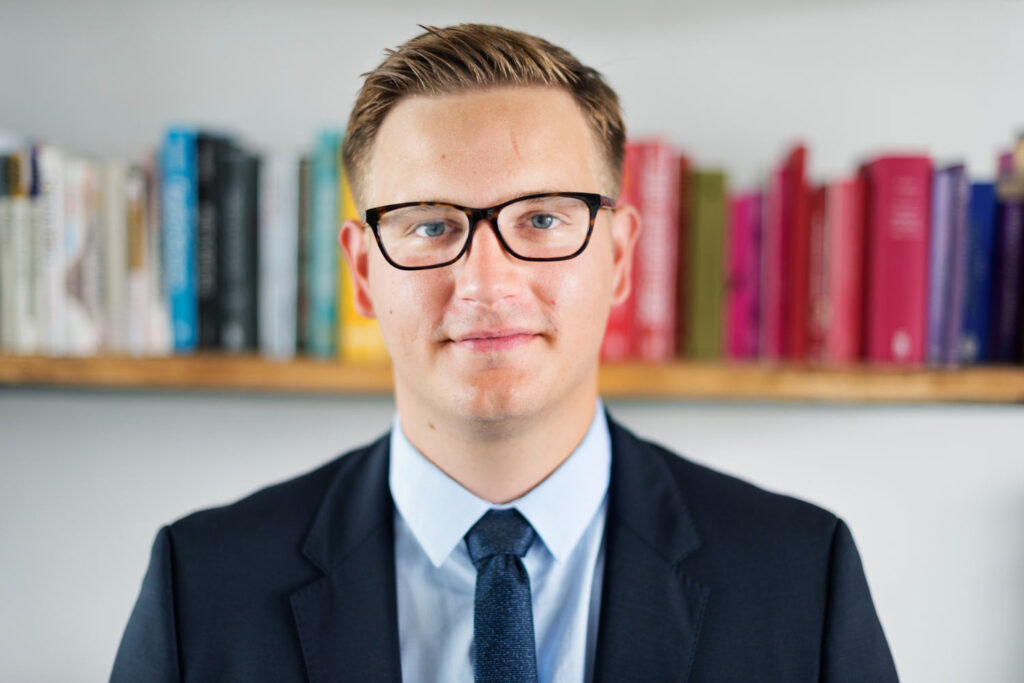 A suited man wearing glasses poses in front of bookshelves, creating a professional library background for a headshot.
