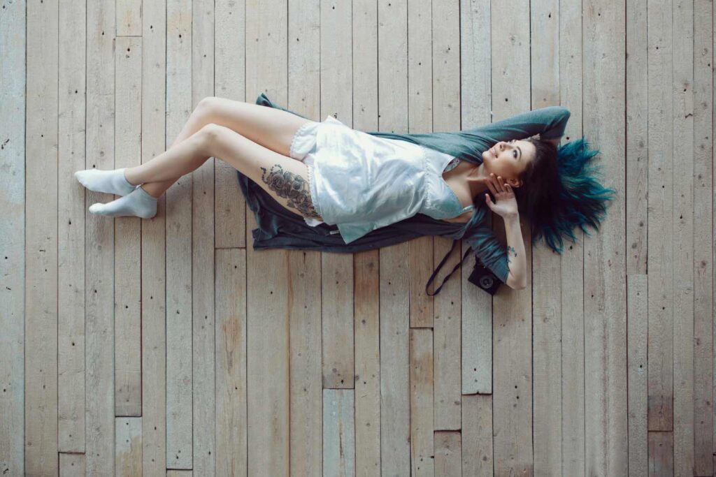 A woman with blue hair rests on a wooden floor, viewed from above, exemplifying top-down lifestyle photography aesthetics.