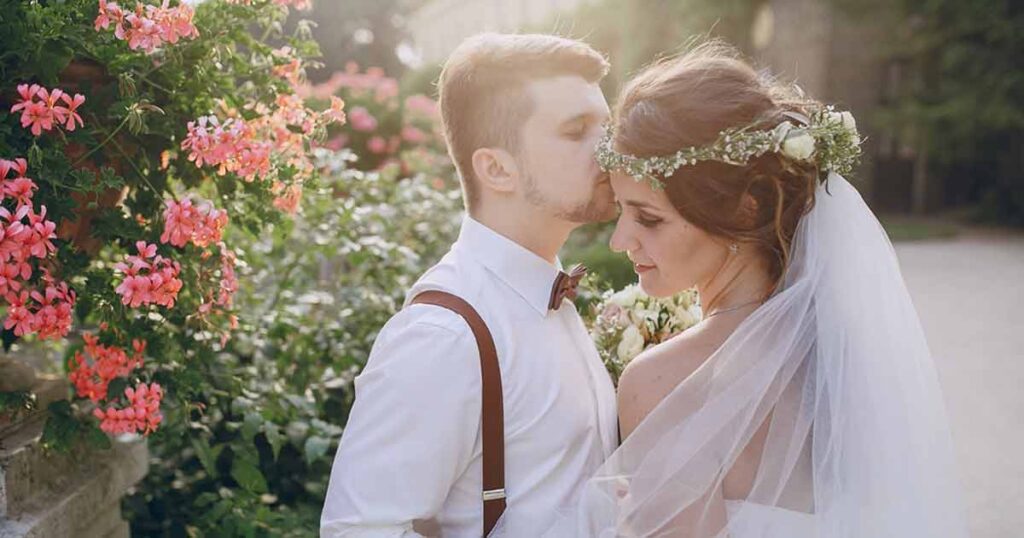 The bride and groom pose in front of colorful flowers, showcasing their love in a beautifully retouched wedding photo.